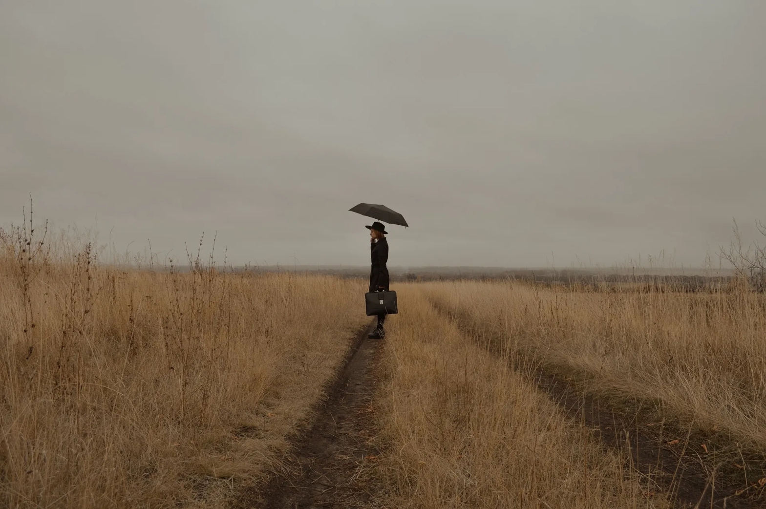 Photograph of a woman standing in the middle of an overgrown, dried out field. She holds a black umbrella over her head, wears a black rimmed hat, a black trench coat, and black boots. She holds a black briefcase. The sky is dark and gloomy.